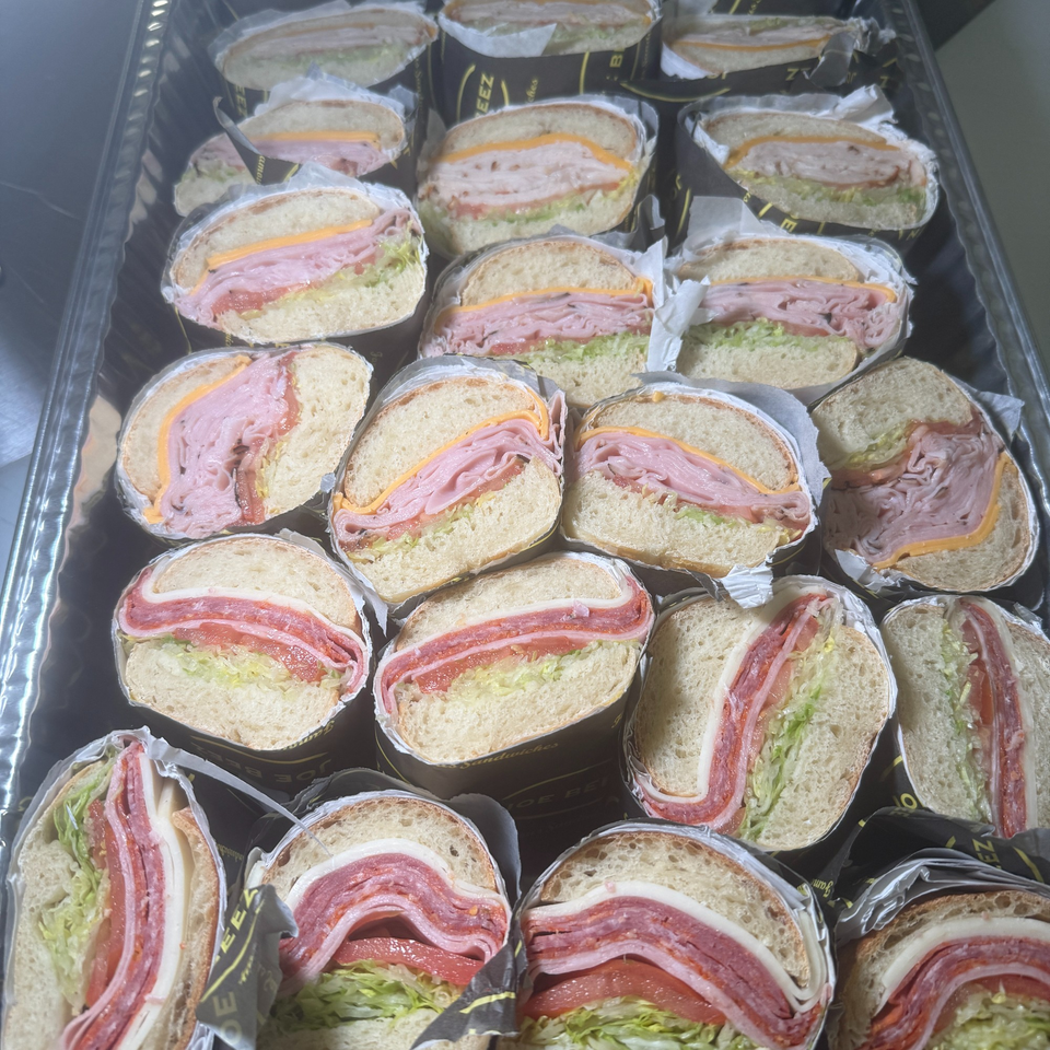 A catering tray filled with individually wrapped sandwiches, some with ham and cheese, others with salami.