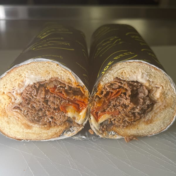 Two halves of a Joe Beez deli sandwich, filled with meat, cheese, and sauce, wrapped in branded paper.