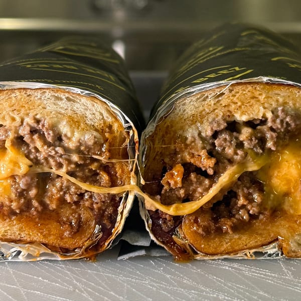 Two halves of a Joe Beez cheesesteak sandwich, wrapped in paper, with melted cheese stretching between them.