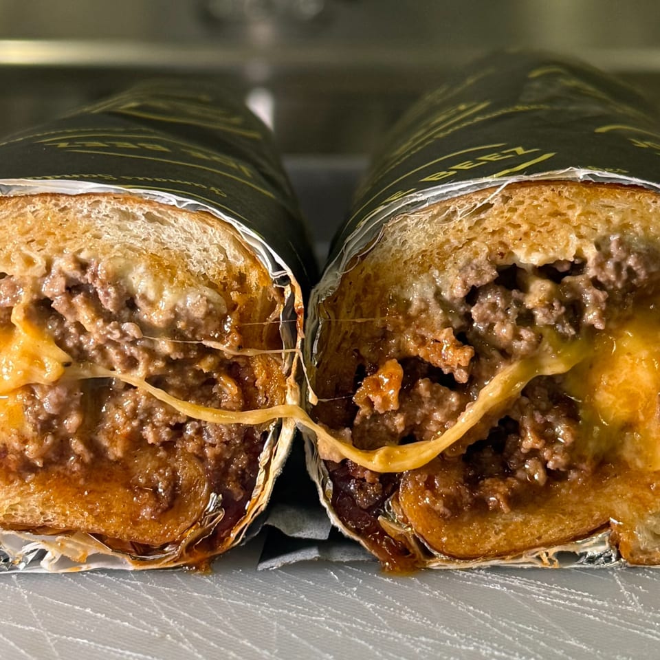 Two halves of a Joe Beez cheesesteak sandwich, wrapped in paper, with melted cheese stretching between them.