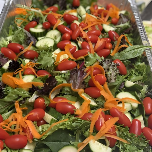 A large catering tray filled with fresh green salad, grape tomatoes, cucumber slices, and carrot shavings.