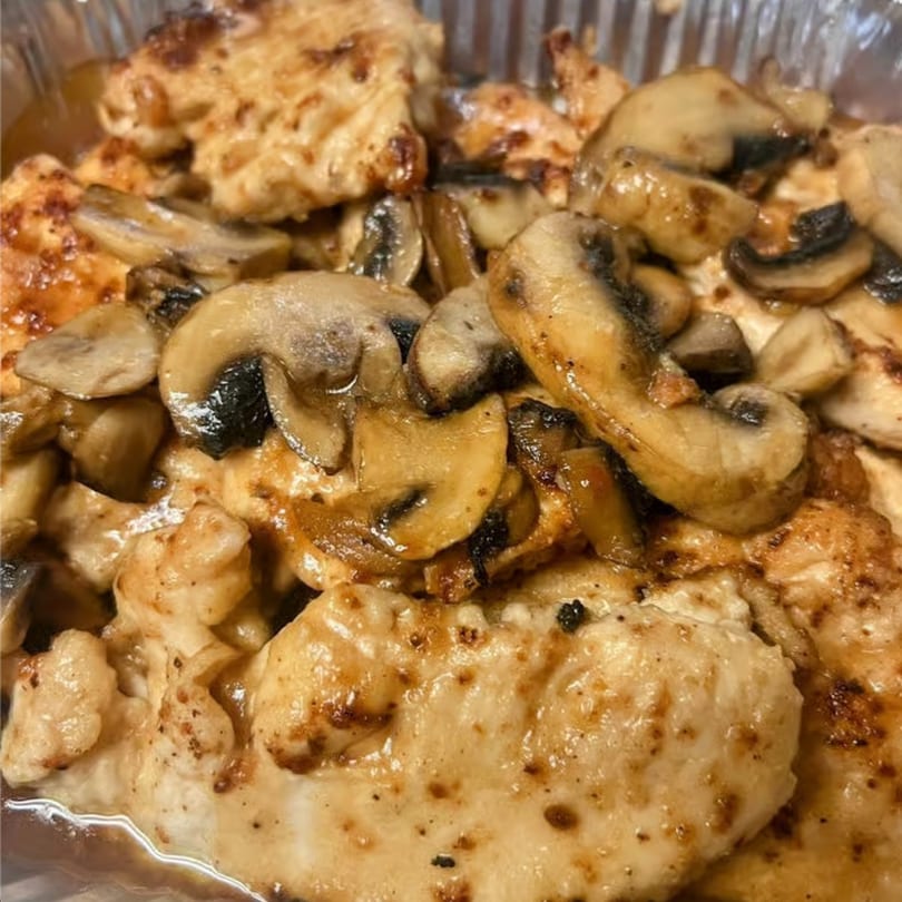 Close-up of golden-brown chicken pieces topped with sautéed sliced mushrooms in a foil container.