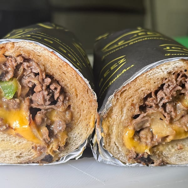 Two Joe Beez deli cheesesteak sandwiches, cut in half, revealing melted cheese, steak, and vegetables.